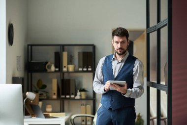 Genç ciddi işadamı tablet kullanıyor ve çalışma masaları ve raflarla birlikte çalışırken kameraya bakıyor.