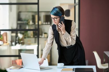 Modern iş dünyasının genç ciddi kadın direktörü cep telefonuyla konuşuyor ve bilgisayarını işyerine göre kullanıyor.