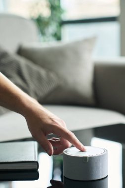 Parlak, modern oturma odasındaki cam sehpadaki akıllı hoparlörleri açmak için uzanan el mobilyaları ve pencerelerden süzülen doğal ışık.