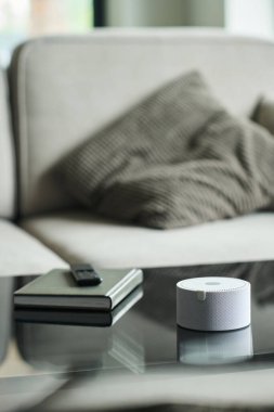 Modern oturma odasındaki cam sehpada oturan ses asistanı cihazı yanında not defteri ve kalemle minimalist ve teknoloji meraklısı bir atmosferi yakalıyor.