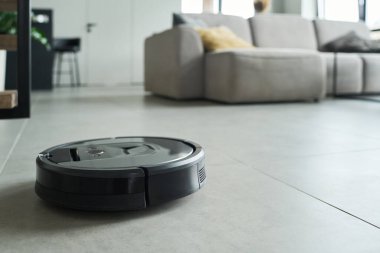 Robotic vacuum cleaner moving across tiled floor of modern living room with beige sofa and large windows visible in background clipart