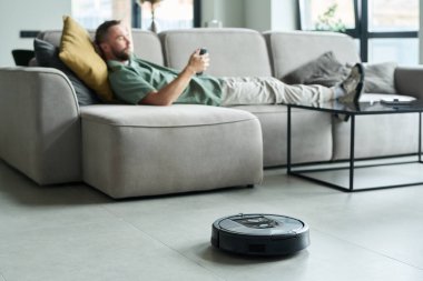 Adam akıllı telefon kullanarak kanepede yatarken robot süpürge de yakınlardaki yerleri temizliyor. Geniş pencereli ve minimalist mobilyalı modern oturma odası.