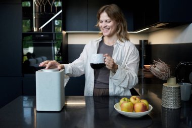 Gülümseyen kadın modern mutfakta elinde kahve fincanı tutarken şık mutfak tezgahında akıllı bir hoparlör kullanıyor.