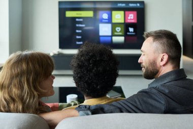 Aile kanepeye oturur, modern oturma odasında sırtları kameraya dönük televizyon izler. Farklı ortam seçenekleri gösteren görünür bir televizyon ekranı