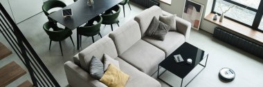 Modern mobilyaları, temiz hatları ve tarafsız tonlarıyla yukarıdan bakan minimalist oturma odası arka planda yemek alanı ve yerde robotik bir vakum oluşturan hoş ve samimi bir atmosfer yaratıyor.