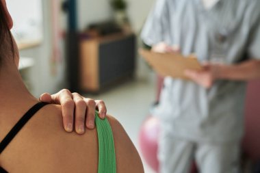 Tıbbi uzman, yeşil giysili bir hastanın omuz muayenesi sırasında pano kullanıyor. Klinik bir ortamda etkileşim gerçekleşiyor