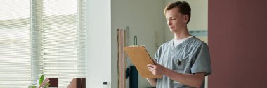 Parlak, modern bir klinikte elinde bir panoyla dikilen, kameradan başka yere bakan, odaklanmış ve içerikte gözüken tıp uzmanları.