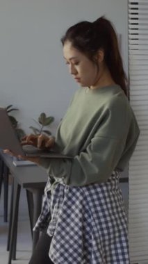 Genç Asyalı bir kadının bilgisayarla çalışırken IT şirketinin kapısına dayandığı orta boy dikey bir fotoğraf, cep telefonuyla konuşan ve etrafta dolaşan bir erkek arkadaş.