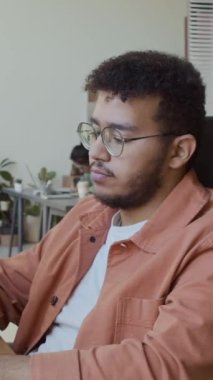 Orta boy dikey mutlu Orta Doğulu genç bir adam. Günlük gömlekli ve gözlüklü. Masasında oturuyor, bilgisayarda çalışıyor, kameraya bakıyor ve gülümsüyor.