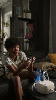 Küçük Afro-Amerikan çocuğun video oyunu oynarken, tıbbi nebulizatörle kanepeye otururken çekilmiş dikey bir fotoğrafı.