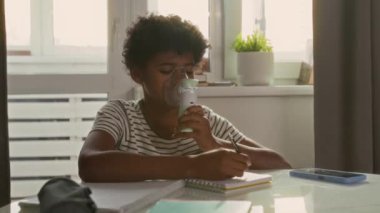 Afro-Amerikan çocuk portresi oturma odasında oturuyor, elinde el yapımı nebulizatör, deftere yazıyor ve kameraya poz veriyor.