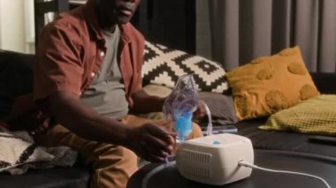 Orta yaşlı siyah bir adam kanepeye yaslanıyor, cep telefonu kullanıyor ve astım ilacını kompresör nebulizörüyle soluyor.