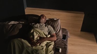 Yatakta uyuyan yaşlı Afro-Amerikalı adamın yüksek açılı görüntüsü, öksürükten uyanması ve evde astım spreyi kullanması.