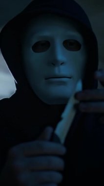 Kapüşonlu isimsiz maskeli bir adamın dikey yakın plan portresi. Keskin bıçağa dokunurken ve kontrol ederken kameraya bakıyor. Yüzünden ve boynundan geçiyor.