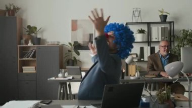 Resmi takım elbise giyen mutlu iş adamı, palyaço peruğu ve burnu açık uzay ofisinde sandalyede dönen, kollarını kaldırıp gülümseyen, yukarıdan dökülen kağıtlar.