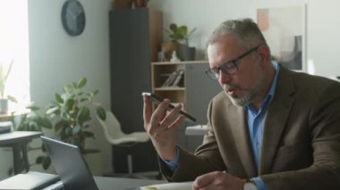 Resmi kıyafetli, olgun gri saçlı iş adamı ofis masasında oturuyor, cep telefonuyla hoparlör modunda konuşuyor ve kağıt üzerinde notlar alıyor.