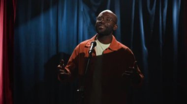 Siyah gözlüklü, parlak turuncu tişörtlü ve beyaz tişörtlü orta boy erkek stand-up komedyeninin hareketli, esprili ve küçük sahnede şarkı söylerken gülüşü.