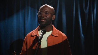Gözlüklü, parlak turuncu tişörtlü ve beyaz tişörtlü siyahi erkek stand-up komedyeninin sahnede dans ederken rastgele hareketler yaptığı ve konuştuğu bir sahne.