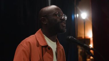 Gözlüklü, beyaz tişörtlü, parlak turuncu tişörtlü, gece kulübünde fıkralar anlatan siyah erkek stand-up komedyenini yakından izleyin.