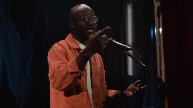 Gözlüklü ve sakallı, beyaz tişörtlü, turuncu tişörtlü siyah erkek stand-up komedyeninin küçük sahnede hareketli ve güvenli bir şekilde mikrofona konuştuğu orta boy pozu.