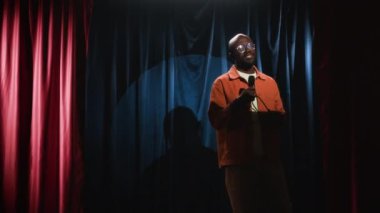Gözlüklü, sakallı, beyaz tişörtlü ve turuncu tişörtlü, sahnede mikrofonla hareket eden ve gelişigüzel yürüyen kendinden emin siyah erkek stand-up komedyeninin orta boy fotoğrafı.