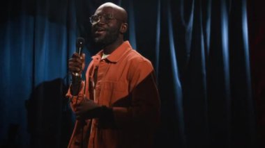Gözlüklü, sakallı, turuncu tişörtlü, siyah erkek stand-up komedyeninin orta ölçekli görüntüsü. Etkin bir şekilde konuşuyorlar ve küçük sahnede mikrofonla yürüyorlar.