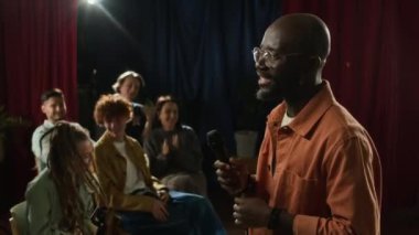 Siyahi erkek komedyenin sakin bir şekilde sahnede konuştuğu ve stand-up gösterisi sırasında salonda oturan bir grup seyirciden alkış aldığı orta ölçekli bir sahne.