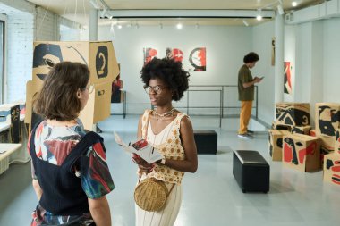 Elinde broşürle genç bir Afrikalı Amerikalı kadın tartışma sırasında başka bir galeri ziyaretçisine bakıyor.