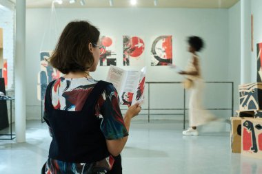 Koyu renk düz saçlı genç bir kadın galeri salonundaki broşürde sergi hakkında bilgi topluyor.