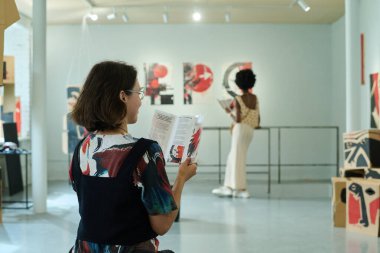 Modern sanat galerisinin genç bayan ziyaretçisinin arka görüntüsü geniş salonda oturuyor ve broşürdeki bilgilere bakıyor.