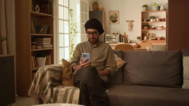 Gözlüklü hoş bir Hintli adam, geleneksel kurta ve türban evdeki kanepede oturuyor ve arkadaşlarıyla cep telefonundan konuşuyor.