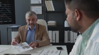 Üst düzey klinik müdürünün modern tıp bürosundaki toplantı sırasında erkek doktorlara çizelgeleri açıklarken omuz üstü fotoğrafı.