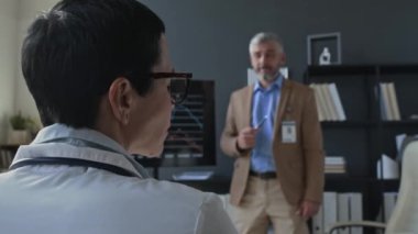 Kadın doktorun ofiste oturduğu, erkek klinik yöneticisinin sunumunu dinlediği ve ofis toplantısı sırasında meslektaşıyla konuştuğu görünümü kapatın.