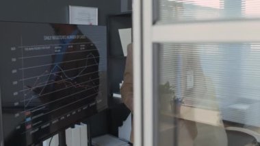 Gri saçlı ve sakallı orta yaşlı bir klinik müdürü, resmi takım elbise giyiyor, ekrandaki veri tablolarına işaret ederken ofis toplantısı sırasında ekibe tıbbi verileri açıklıyor.