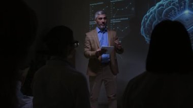 Yenilikçi sinirsel implant teknolojisini sunan sağlık uzmanı, karanlık konferans odasındaki tıp uzmanlarına ekranda beyin ve veri eğilimlerinin görsel projeksiyonlarını sunuyor.