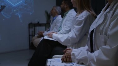 Beyaz önlüklü bir grup sağlık uzmanı karanlık konferans odasında oturmuş konuşmacı ve alkışları dinliyorlar.