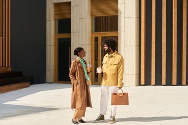 Afrika kökenli Amerikalı erkek ve siyah kadın, güneşli havalarda yürürken modern ofis binasında ellerinde kahve fincanları ve evrak çantasıyla sohbet ediyorlar.