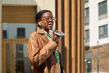 Modern binanın dışında konuşurken ve yürürken akıllı telefonu yüzüne yakın tutan bir kadın. Kahverengi bir ceket ve gözlük takıyor, kendinden emin ve odaklanmış görünüyor.