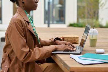 Bilgisayarı kullanan kişi arka planda modern ofis binaları ve işe odaklanmanın yanında bir de içki olan açık hava ahşap bir masada oturuyor.