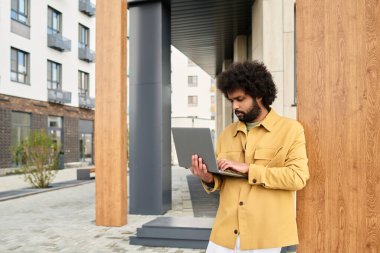 Modern apartmanların yanında kaldırımda dikilirken dizüstü bilgisayar kullanan afro saçlı bir adam. Dekor çağdaş mimari ve ahşap sütunları içeriyor