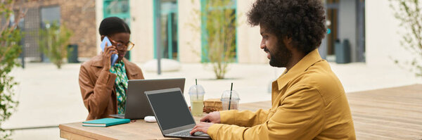 Коллеги, работающие за открытым столом с ноутбуками и напитками, ведут беседу. Один коллега многозадачность при использовании телефона и носить очки