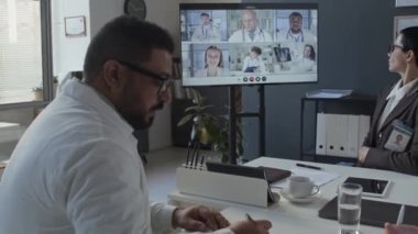 Ekranda gösterilen web konferansında meslektaşlarını dinlerken ve ofis masasındaki not defterine not alırken çok ırklı doktor pozunu aşağı eğ.
