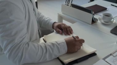 Beyaz önlüklü ve gözlüklü erkek doktorların ofiste ekranda gösterilen çevrimiçi video konferansında meslektaşlarının sunumlarını dinlerken not alırken görüntülerini yukarı kaldır.