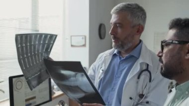 Orta yaşlı Kafkasyalı doktor ve çok kültürlü erkek meslektaşı tıbbi ofiste yan yana duruyor, röntgenleri inceliyor ve iş günlerinde tartışıyorlar.