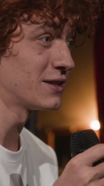 Beyaz tişörtlü, beyaz saçlı, gülümseyen beyaz erkek stand-up sanatçısının sahnedeki performansını ve seyircilere bakıp mikrofona fıkralar anlattığı dikey yakın çekim.