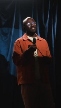 Gözlüklü, sakallı, beyaz tişörtlü ve turuncu tişörtlü, sahne performansı sırasında mikrofonla hareket eden ve gelişigüzel yürüyen kendine güvenen siyah erkek stand-up komedyeninin dikey görüntüsü.