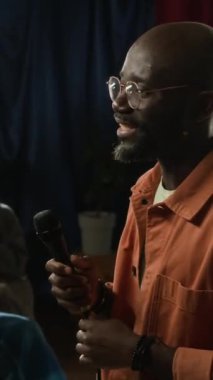 Siyahi erkek komedyenin sakin bir şekilde sahnede konuştuğu ve stand-up gösterisi sırasında salonda oturan izleyicilerden alkış aldığı dikey çekimler.