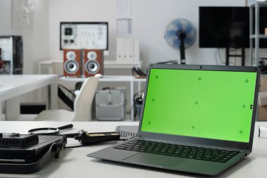 Laptop with green screen and various electronic devices in modern home office. Background shows speakers, fan, and other equipment clipart