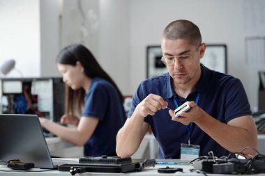 Bir teknoloji laboratuvarında küçük bir elektronik bileşeni incelemeye odaklanan bir adam. Arka planda çalışan kadın, her ikisi de teknik ekipman içeren görevlerle meşgul.