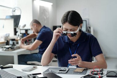 Laboratuvar önlüklü genç teknisyen büyüteç kullanarak akıllı telefon parçalarını inceliyor. Masada oturan teknisyen, arka planda çalışan başka bir teknisyenle birlikte araçlarla çevrili.